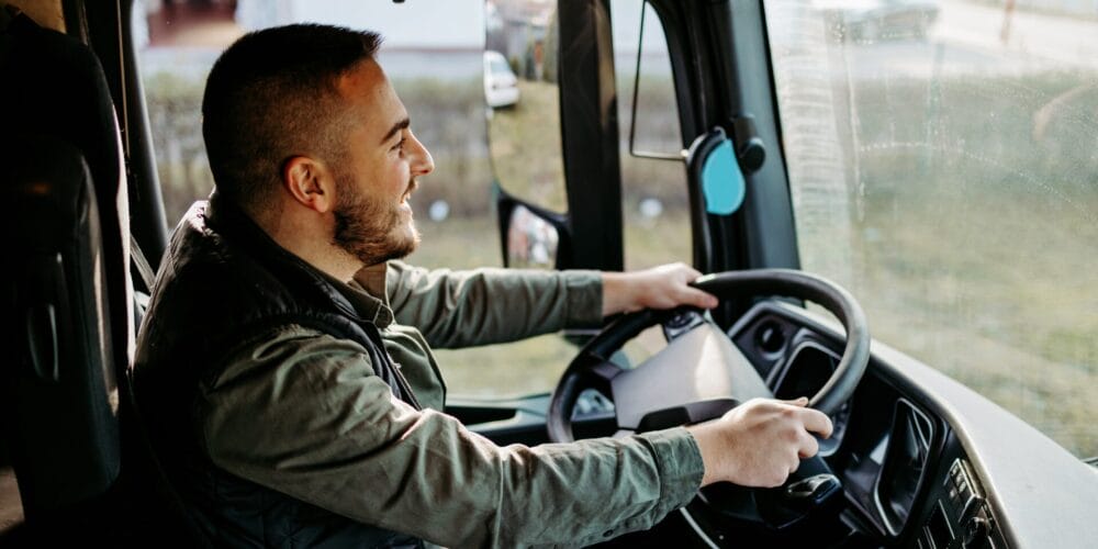 Smiling truck driver holding the steering wheel while driving a commercial vehicle during the day.