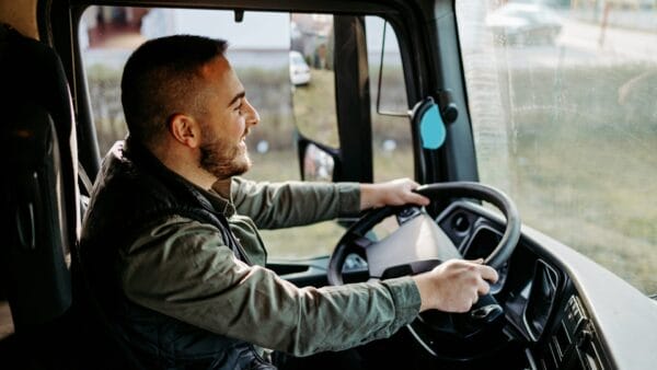 Smiling truck driver holding the steering wheel while driving a commercial vehicle during the day.