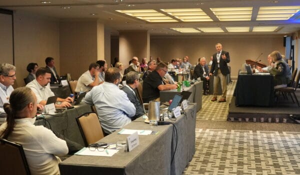 A professional conference room filled with attendees seated at long tables, listening to a speaker standing and presenting near the front. Participants are using laptops and taking notes, while nameplates and beverages are visible on the tables. The setting suggests a formal industry meeting or council session, with a collaborative and engaged audience.