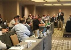 NMFTA Fall Meeting 2025 A professional conference room filled with attendees seated at long tables, listening to a speaker standing and presenting near the front. Participants are using laptops and taking notes, while nameplates and beverages are visible on the tables. The setting suggests a formal industry meeting or council session, with a collaborative and engaged audience.